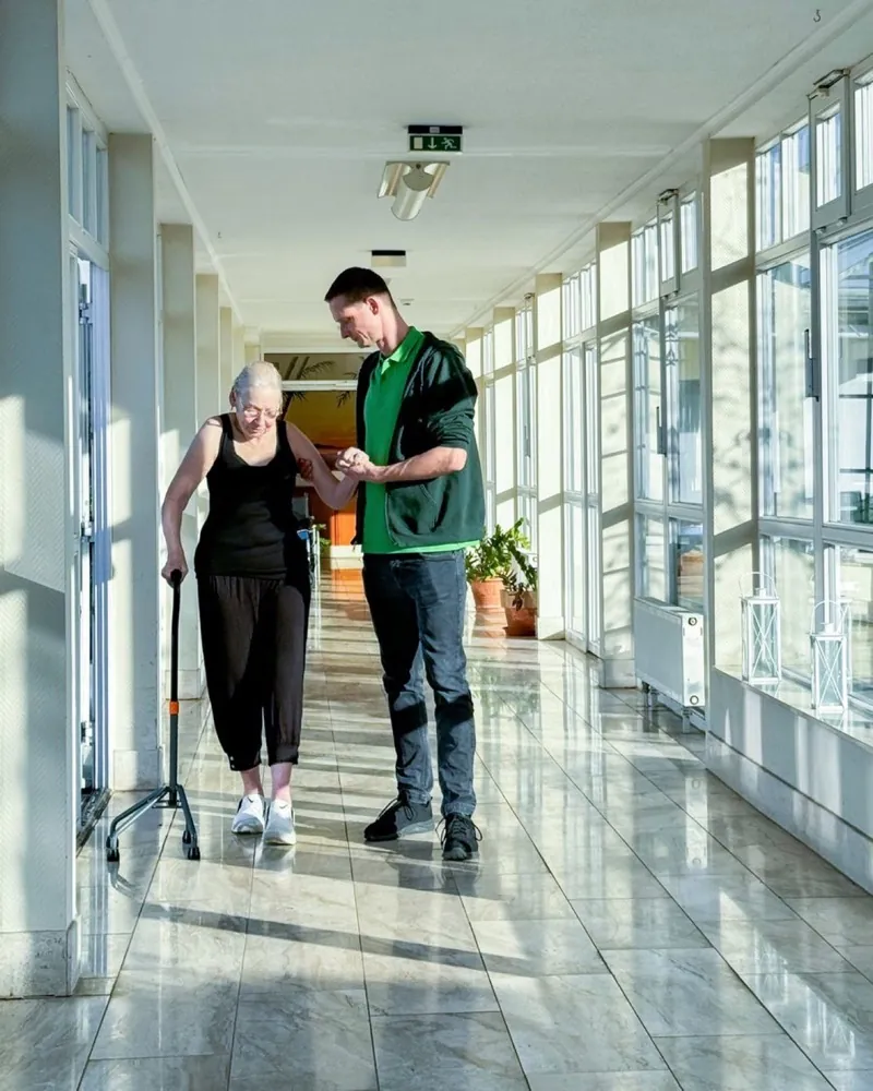 Therapeut begleitet eine Bewohnerin mit Gehstock durch den Flur einer Pflegeeinrichtung