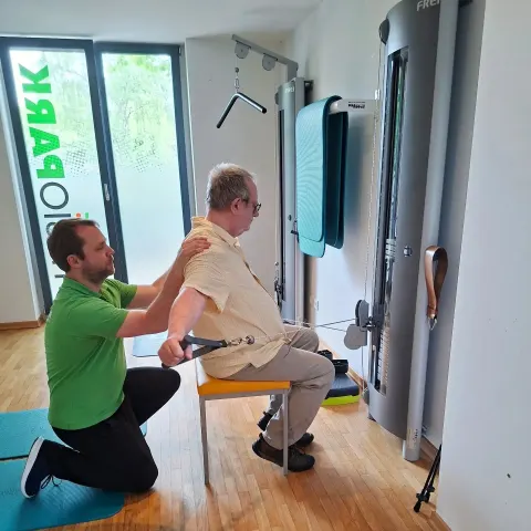 Therapeut stabilisiert die Schulter eines Patienten beim Seilzugtraining in der Praxis Tempelhof