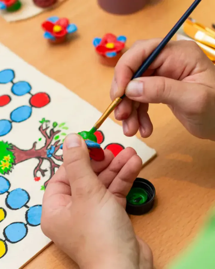 Hände bemalen eine kleine Figur, daneben ein bemaltes Spielbrett und Pinsel