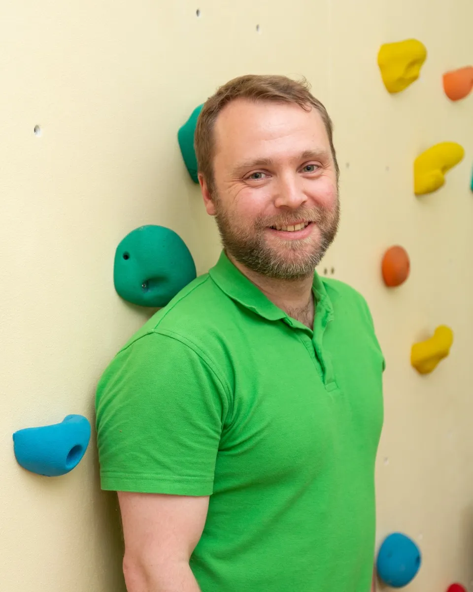 Therapeut im grünen Poloshirt lacht vor der Kletterwand der Praxis Tempelhof